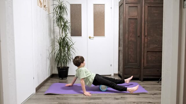 Caucasian child boy rolling the roller on legs making self-massage. Doing a fascia massage lying on yoga mat, wearing a sports clothes at home. European sportsman is training legs. Morning routine. 4K