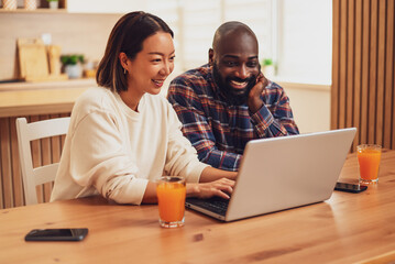 A cheerful couple sits together at a wooden table, smiling and laughing while looking at a laptop...