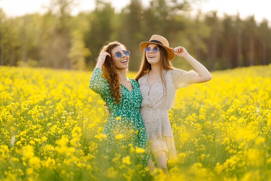 Two cheerful friends in dresses are having fun in a blooming field, bathed in sunlight. The young women pose outdoors, enjoying nature. Concept: style, fashion, and friendship.