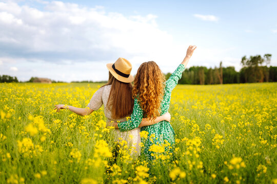 Two cheerful friends in dresses are having fun in a blooming field, bathed in sunlight. The young women pose outdoors, enjoying nature. Concept: style, fashion, and friendship.