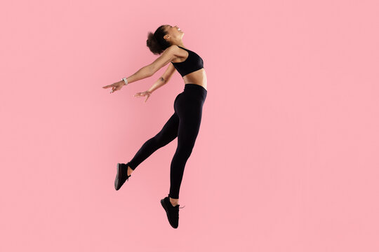 Athletic black woman jumping backward with arms open on pink studio background, expressing freedom, energy and dynamic movement in fitness lifestyle concept