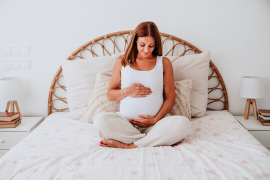 Pregnant woman sitting on bed at home touching belly in cozy bedroom