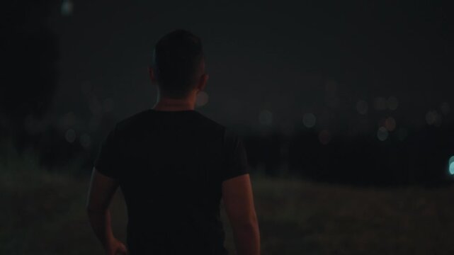 Cinematic footage of a man standing alone overlooking city lights at night. Urban night scene with soft bokeh lights, representing solitude, reflection, and emotional mood in a modern city environment