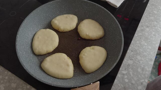 A woman hand lifts a glass lid from a frying pan where pancakes are cooking.