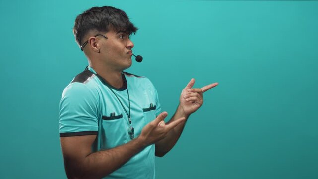 Man referee in turquoise uniform pointing fingers and pursing lips with headset microphone visible in studio; playful doubt.