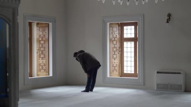 Man performs prayer in empty room near window, ritual movement, spirituality.
