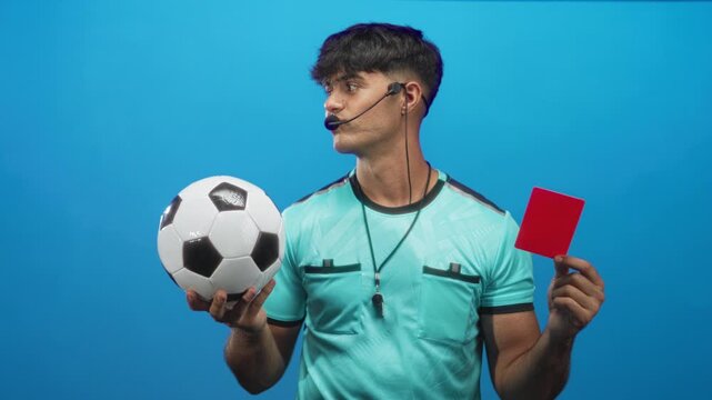 Man referee holding soccer ball and showing red card with headset and whistle in studio; authority discipline.