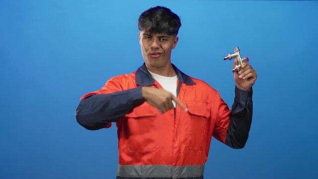 Man plumber wearing orange coveralls holding chrome tap and points finger to tap in studio; friendly repair service.