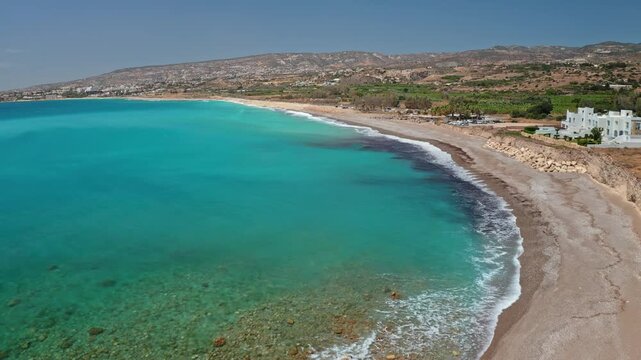 Cyprus coastline with beautiful turquoise Mediterranean sea meeting a long pebble beach, with a white building on the rocky shore under a clear summer sky during a sunny day. Aerial view drone flight