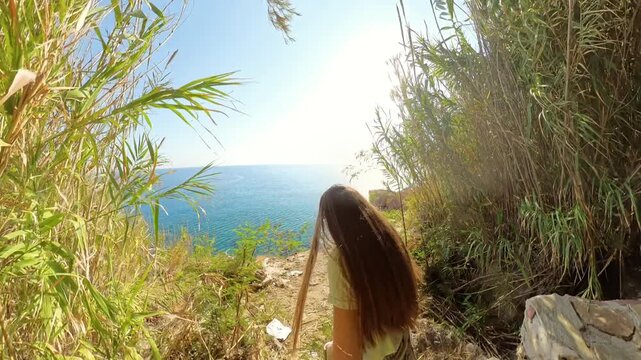 A woman walks along a rustic stone pathway toward a cliff overlooking the sea, framed by dense foliage with sunlight filtering through. The scene evokes a sense of adventure and tranquility by the oce