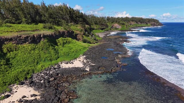 Keyframe 1: Aerial shot of tropical coastline. Blue ocean meets black lava rocks. Waves crash along the shore. Green cliffs rise in the background. Sunny day highlights natural colors