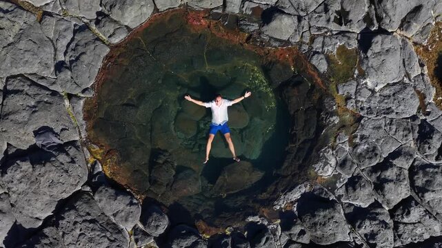 Aerial view shows a person lying in a rock pool with arms outstretched. Relaxed expression reflects deep contentment. Surrounding landscape includes green forests, ocean, and sand