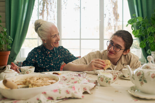 Senior Caucasian woman smiling while sitting beside teenage Caucasian boy eating pastry at table set with teapot and dishes, both engaging in warm interaction near window with green curtains