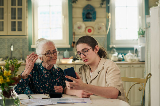 Senior Caucasian woman sitting beside teenage Caucasian boy showing smartphone screen, both focusing on device, paperwork and calculator on table in bright kitchen setting