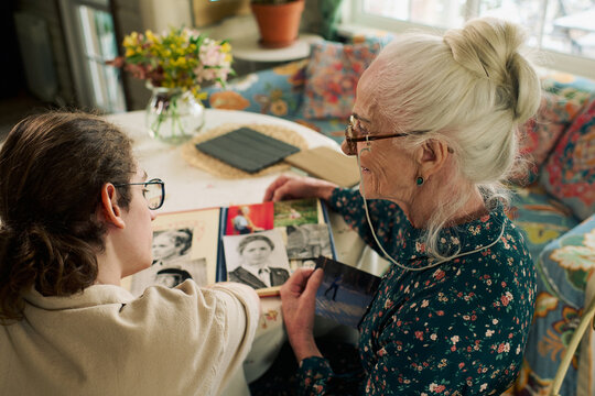 Senior Caucasian woman and young adult Caucasian man sitting together at table reviewing old family photographs, both wearing glasses, engaging in conversation and reminiscing