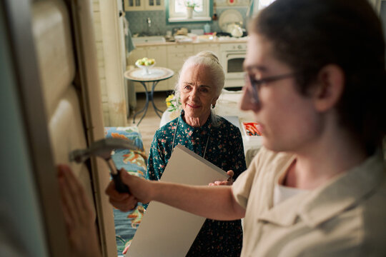 Senior Caucasian woman standing and smiling while young adult Caucasian man with long hair using hammer to drive nail into wall in home kitchen, both focusing on household task together
