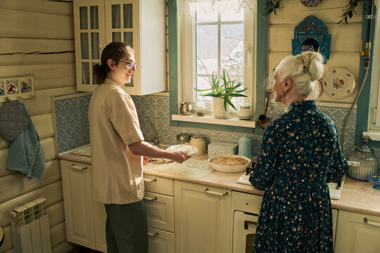 Caucasian young adult man smiling while holding plate and talking with Caucasian senior woman in rustic kitchen, both standing near counter with homemade baked goods and window in background