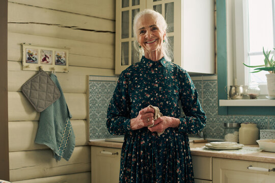 Portrait of senior Caucasian woman smiling while standing in rustic kitchen holding dish towel, showing long gray hair and wearing floral dress, natural light coming from window