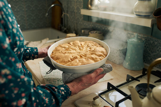 Senior Caucasian woman holding freshly baked pie in kitchen, steam rising from hot dish, standing near stove with hands visible, wearing patterned long sleeve shirt, preparing homemade dessert