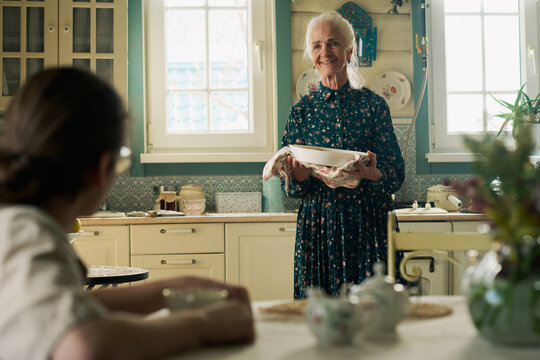Senior Caucasian woman smiling while holding baking dish in vintage kitchen, interacting with young adult Caucasian man sitting at table, sharing homemade meal