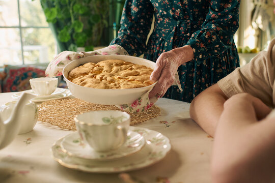 Senior Caucasian woman serving freshly baked pie at dining table with young adult Caucasian man sitting nearby, hands visible, vintage porcelain tea set arranged on table