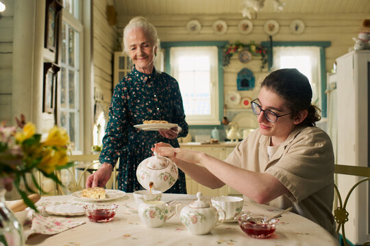 Senior Caucasian woman smiling while standing beside young adult Caucasian man pouring tea into floral teacup at table with teapot, cups, and plates in cozy kitchen setting