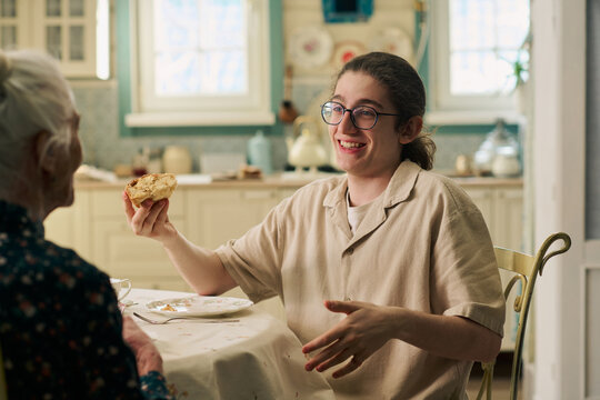 Caucasian teenager boy with long hair and glasses smiling while holding piece of pie, sitting at table with senior Caucasian woman in kitchen, engaging in friendly conversation