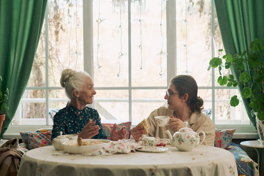 Senior Caucasian woman smiling and talking with young adult Caucasian man sitting at round table sharing tea and pastries in front of large window with green curtains