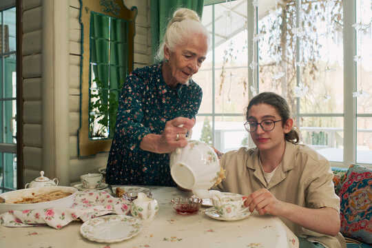 Senior Caucasian woman pouring tea for teenage Caucasian boy sitting at table, both engaging in traditional tea time ritual with porcelain teapot and cups in bright indoor setting