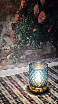 Candlelit Tranquility in Bloom: A softly lit candle in an ornate blue glass holder casts a warm glow, set against the backdrop of a blossoming rose bush.