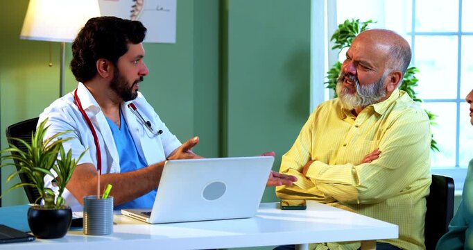 Professional Indian young male doctor using laptop computer with Asian senior retired couple patients across a table in a modern clinic; healthcare, digital technology, and old age care concept.