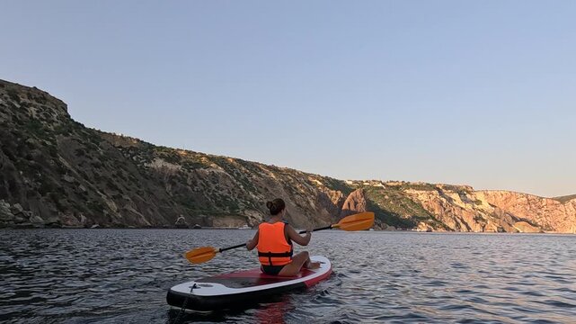 Paddleboarding, woman, sea adventure. Young woman in life vest paddles a stand-up paddleboard on calm ocean water near majestic coastal cliffs at sunset.