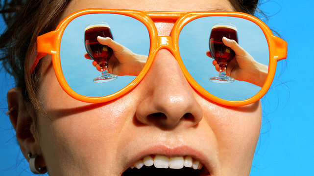 Woman in orange sunglasses with dark beer reflected in lenses under blue sky. Concept of craft beer advertising, premium beverage campaign, product lifestyle visuals, summer promotion, bar marketing.
