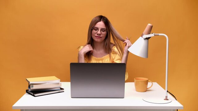 Playful woman working on laptop playing with her hair while sitting at a desk with a yellow background