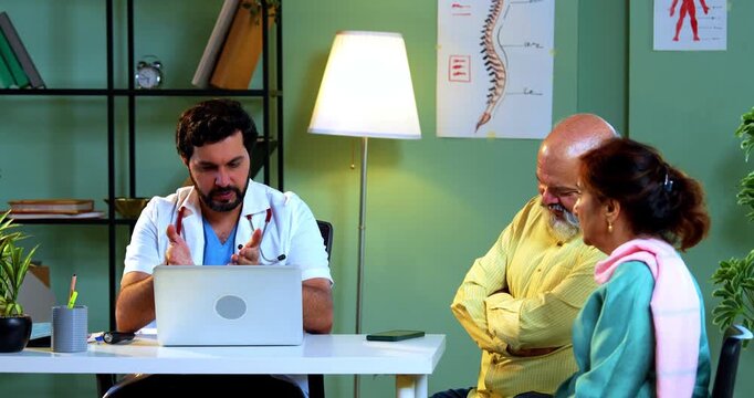 Professional Indian young male doctor using laptop computer with Asian senior retired couple patients across a table in a modern clinic; healthcare, digital technology, and old age care concept.