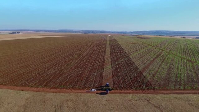 Tractor with precision planter operating along soybean field in Formosa, Goi&aacute;s, Central-West Brazil, with visible planted rows, field sections and support vehicle on dirt road, drone pulling out shot.