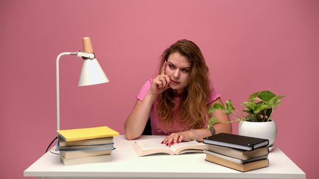 Stressed college student struggling with exam preparation and yawning while studying a textbook on a pink background