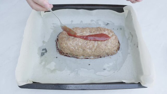 Hand spreads tomato sauce over cooked meatloaf on baking tray from top view