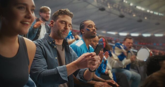 Stadium Championship Match: Man Using Smartphone for Online Sports Betting. White Male Fan Checks Odds in the Stands, Then Celebrates Loudly as His Team Scores During the Packed League Game.