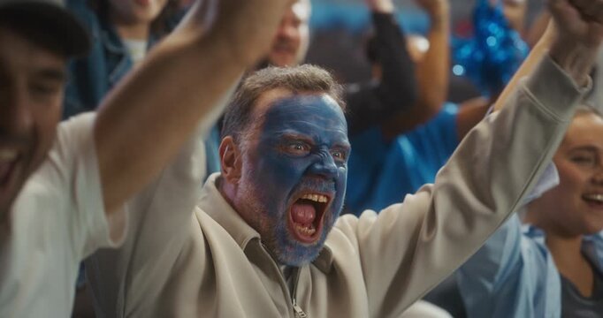 Close-up Portrait of a White Male Soccer Fan With Blue Painted Face Roaring in Celebration, Arms Raised Among Smiling Supporters at a Packed Stadium During a Pro League World Tournament Match.