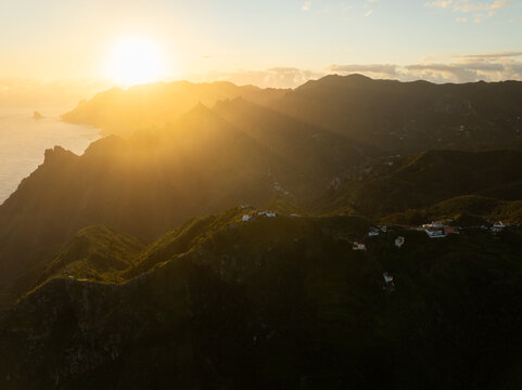 Aerial view of the sun's radiant fingers stretch across the rugged terrain, illuminating the scattered homes nestled among the peaks, Las Carboneras, Canarias, Spain.