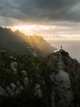 Aerial view of sun rays piercing through the dramatic clouds over the rugged mountains and the vast ocean, a lone figure standing atop a peak, Santa Cruz de Tenerife, Canarias, Spain.