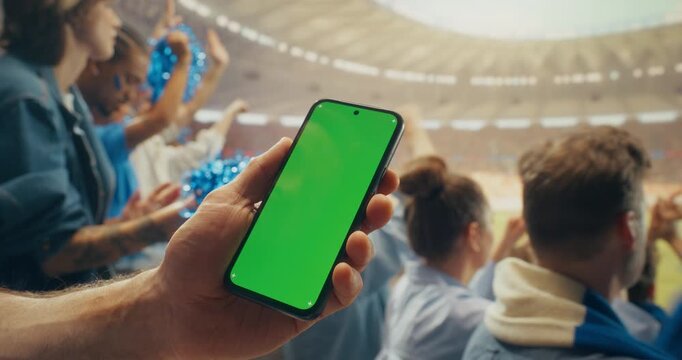 Close-up of Hand Holding a Green Screen Smartphone Mockup During a Packed Soccer Championship in Stadium With Cheering Fans, Raised Arms. Betting App and Screen Insert Concept.