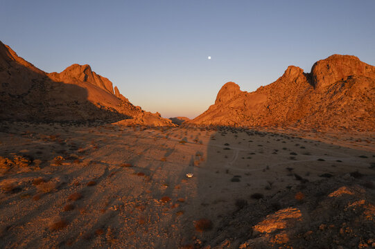 Aerial view of rugged, sun-kissed peaks and valleys cast long shadows over the arid landscape, a solitary vehicle traversing the dusty terrain, Spitzkoppe, Namibia.