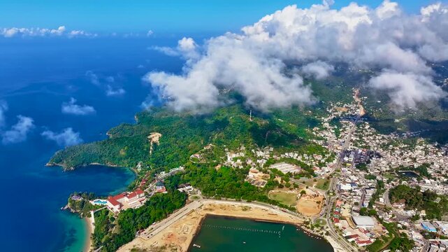 An aerial view of fluffy white clouds over a modern coastal city. Cloud formation meets clear skies over a cityscape. Santa Barbara de Samana, Dominican Republic, from a bird's eye view.