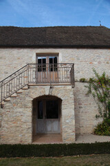 Stone building exterior featuring a wooden door, wrought iron balcony, and staircase surrounded by greenery in a rural French setting