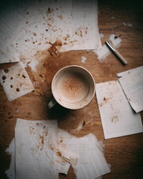 Spilled coffee cup on wooden desk with scattered papers and pen. Home office, natural mess, overhead shot
