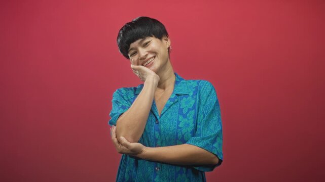 Woman with short black haircut wearing blue patterned shirt, touching cheek with hand while supporting elbow with other arm, smiling in studio; joyful.