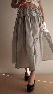 A young woman with a brunette bob hairstyle showcases a stylish two-piece outfit featuring a white open-back crop top and a high-waisted, tiered gingham check maxi skirt in a minimalist indoor setting