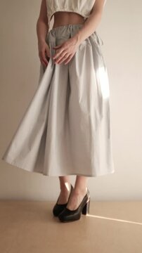 A young woman with a brunette bob hairstyle showcases a stylish two-piece outfit featuring a white open-back crop top and a high-waisted, tiered gingham check maxi skirt in a minimalist indoor setting